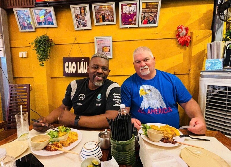 Two men dining at a restaurant with yellow walls, posters, and a menu board. 1