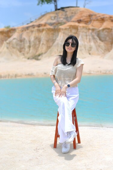 A person sitting on a wooden chair by a beach with clear blue water and a rocky cliff in the background. 2