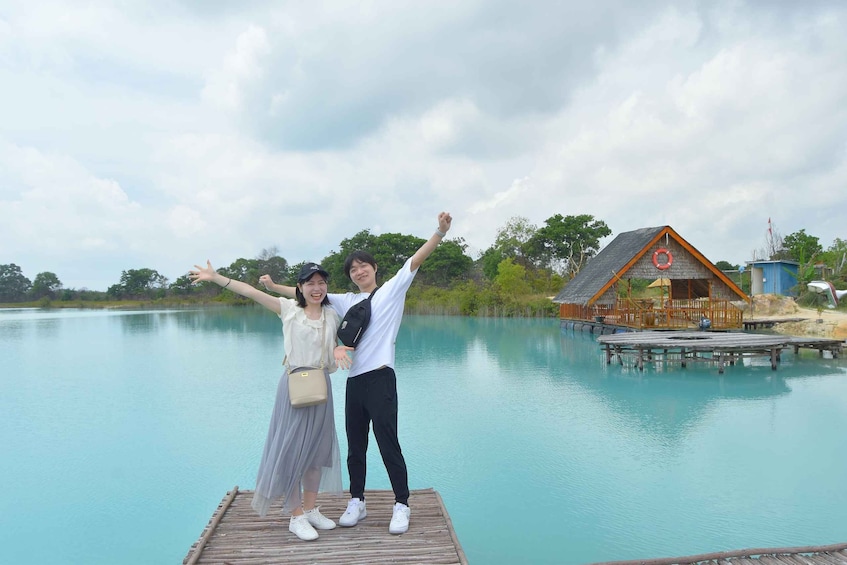 Two people standing on a wooden dock by a calm lake, with a wooden house and dock in the background. 3
