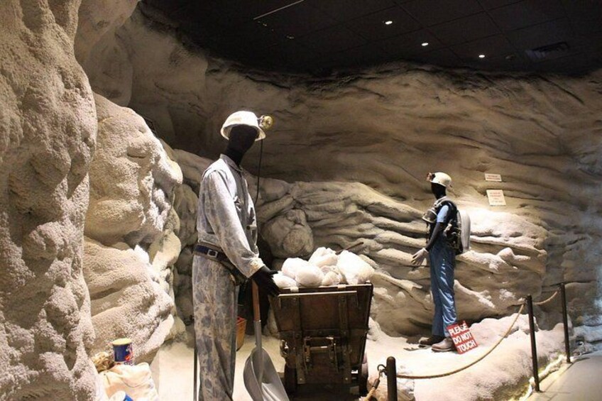 En museiutställning med två skyltdockor klädda i gruvarbetarkläder, en vagn med gruvarbetarverktyg och en bergig bakgrund som liknar en grotta. 4