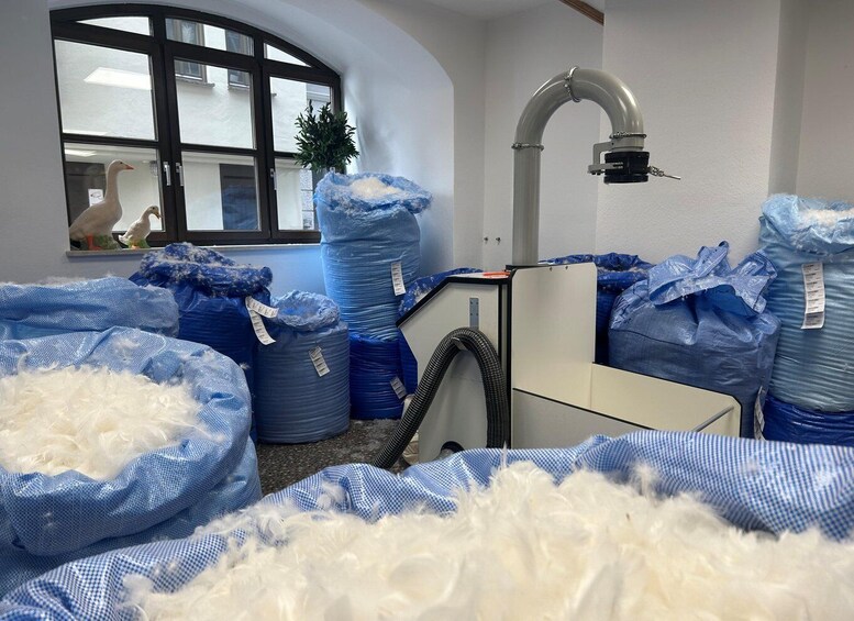 A room with large blue bags filled with white feathers, a window with a view of a building, and a curved pipe on the wall. 5