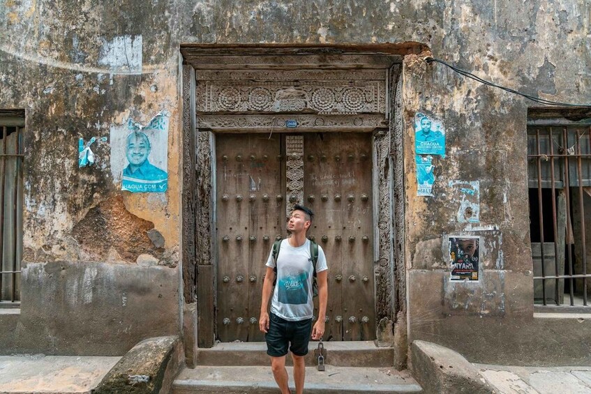 A person standing in front of a weathered building with a large wooden door and decorative carvings. 3