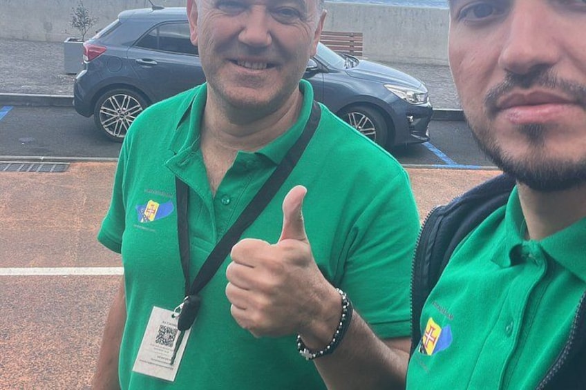 Two individuals in green shirts with logos, one giving a thumbs-up, standing outdoors with a car and a building in the background. 3