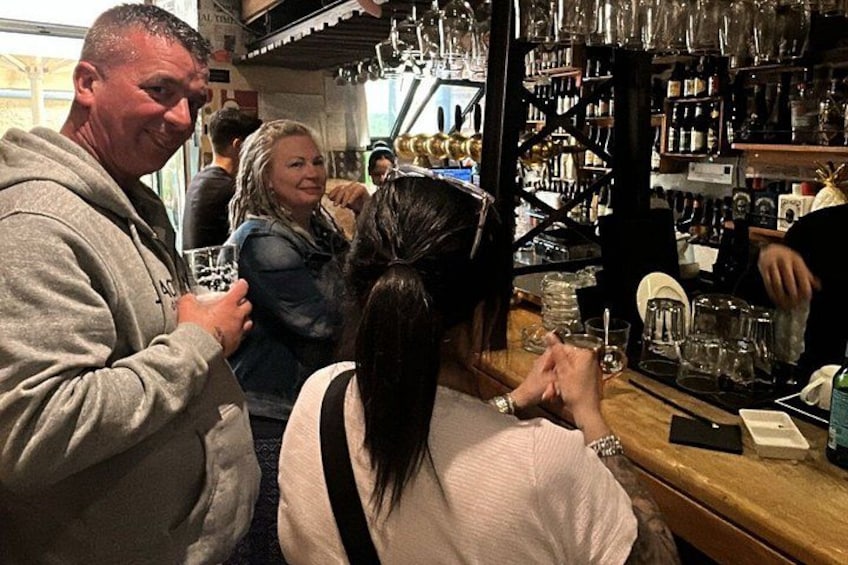 A bar with people standing and conversing, shelves stocked with bottles, and a counter with glasses. 2
