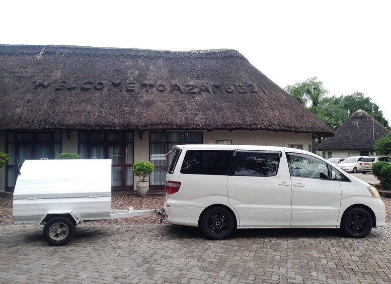 A white van parked in front of a building with a thatched roof and the words "WELCOME TO AZAMGEEZ" carved into it. 2