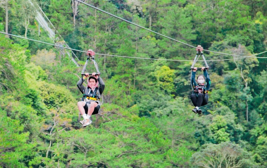 Two people suspended in mid-air on a zipline over a forested area. 1