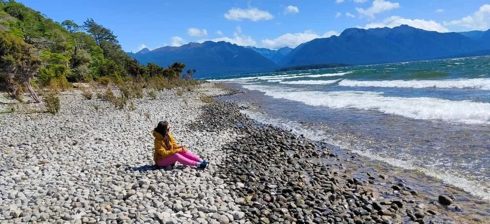 A person sitting on a pebble beach with a mountain range in the background. 1