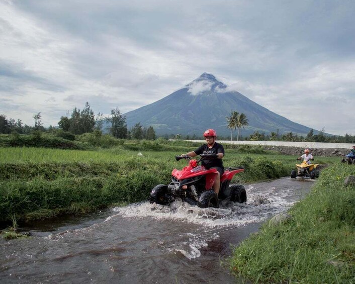 A person riding a red quad bike through a shallow stream with a volcano in the background. 1