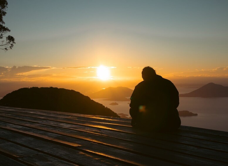 A person sitting on a wooden bench overlooking a sunset over a body of water with distant landforms. 1
