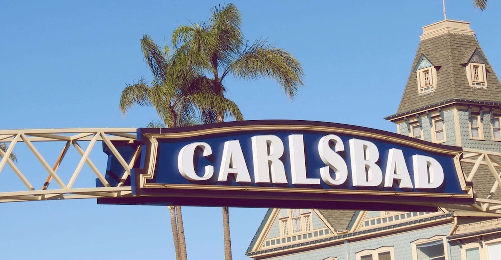 A sign with the word 'CARLSBAD' against a clear blue sky, with palm trees and a building in the background. 1