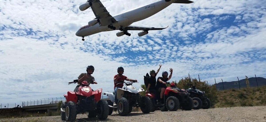 Four people on quad bikes, a large airplane flying overhead, and a fence in the background. 5