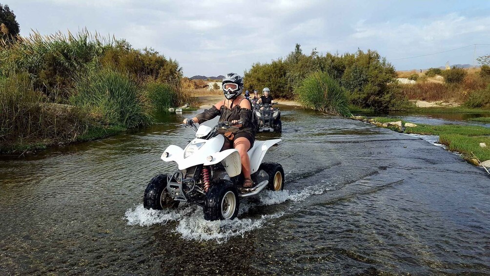 A person riding a white ATV on a narrow waterway surrounded by vegetation. 1
