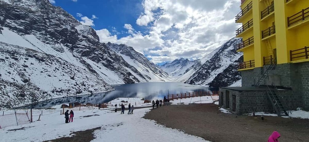 A snow-covered mountain landscape with a bridge over a lake, a yellow building, and people walking. 5