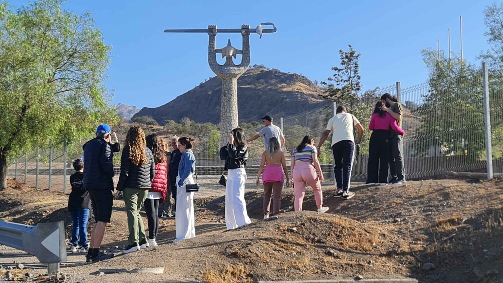 A group of people standing near a large stone structure with a crossbar and weights, surrounded by a fenced area with trees and a mountain in the background. 2
