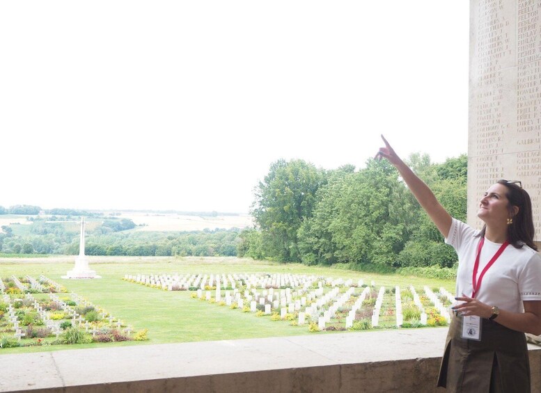 A person pointing at a memorial with rows of white crosses, a white monument, and a green landscape in the background. 1