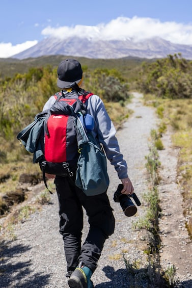 A person walking on a dirt path with a backpack and camera, with a mountain in the background. 1