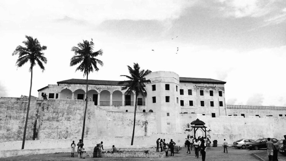 A historical building with arched windows and palm trees, surrounded by a wall and a few people. 1