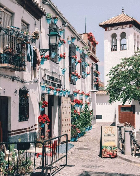 A narrow street lined with white buildings adorned with blue flower pots and red flowers. A signboard for Marrakech is visible. 5