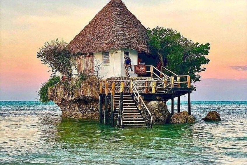 A thatched-roof house on stilts over water with a sign reading "The Rock". 1