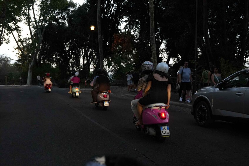 People riding scooters on a road lined with trees. 3