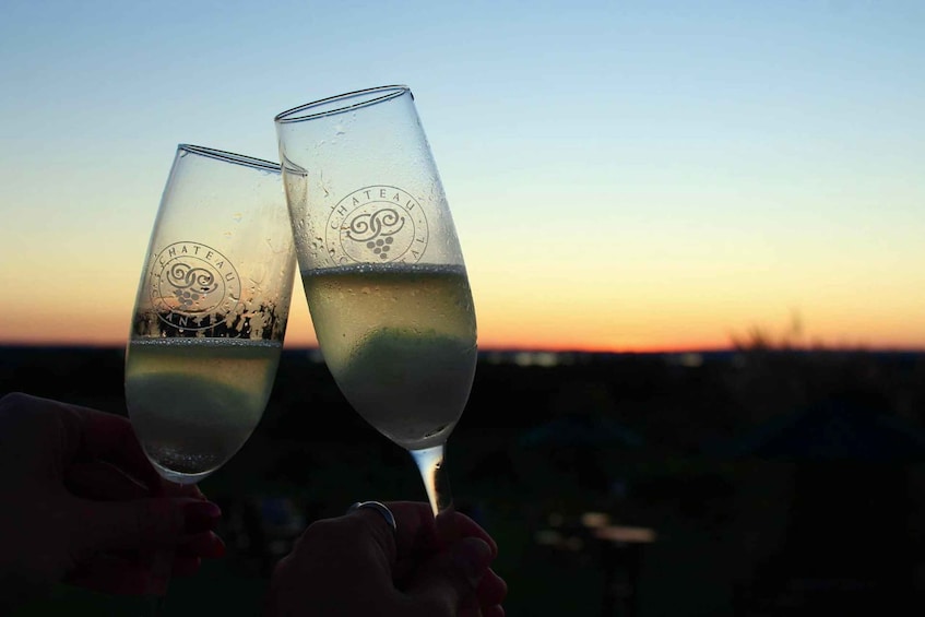 Two glasses of white wine with 'CHATEAU' embossed on them, held by hands against a sunset background. 1