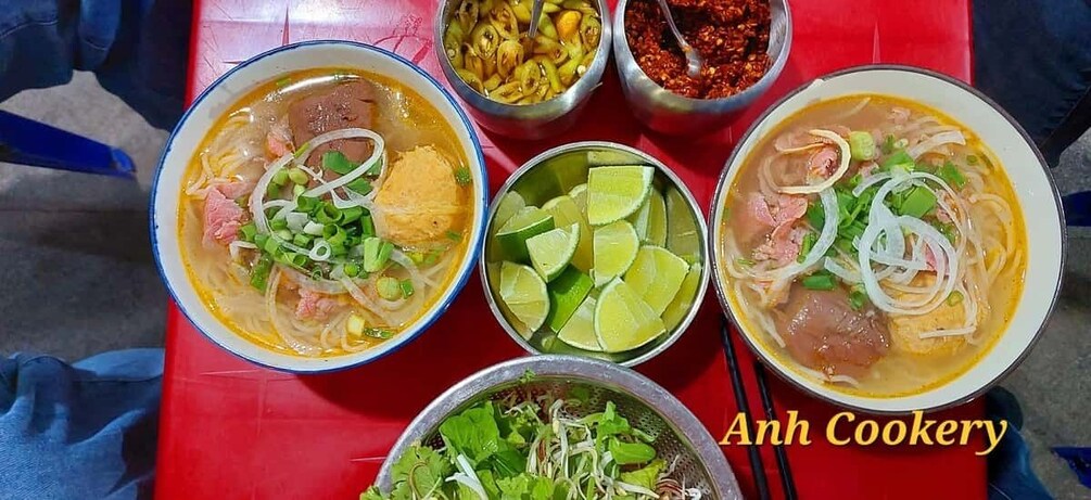Three bowls of soup with various toppings, a bowl of sliced lime, and a bowl of greens on a red table. 3