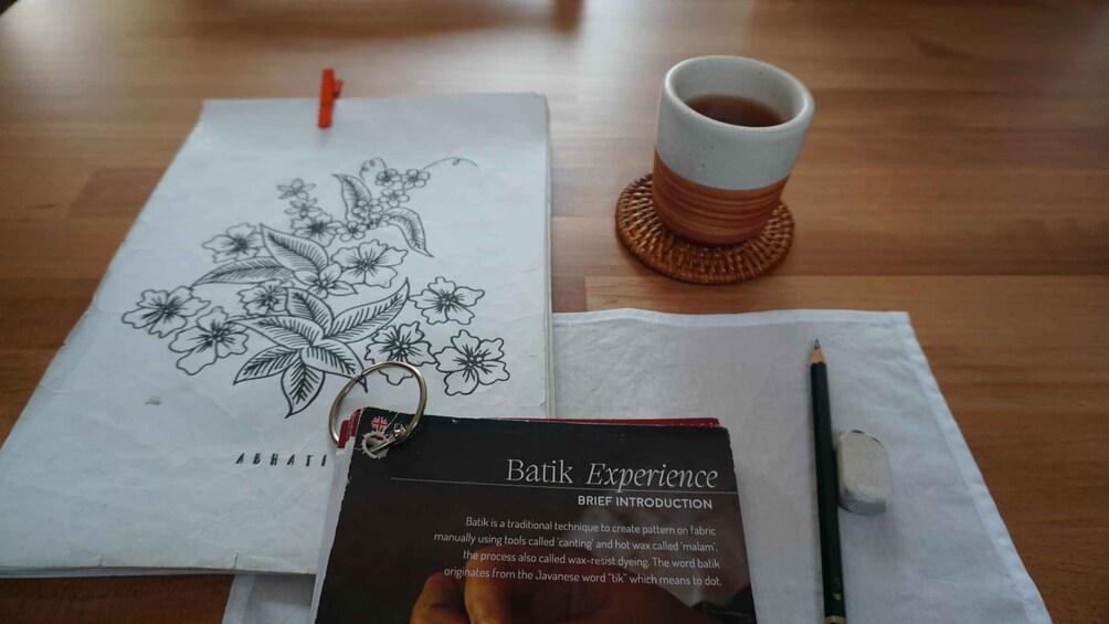 A notebook with a floral drawing, a cup of coffee, a pencil, and a booklet titled 'Batik Experience'. 4