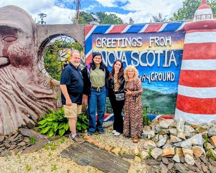 A group of people standing in front of a colorful mural with the text 'Greetings from Nova Scotia' and a lighthouse. 1