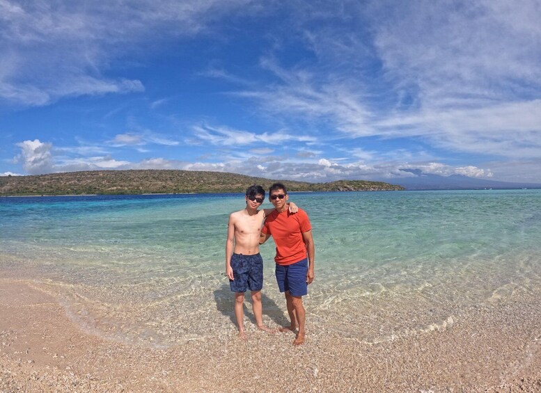 Two people standing on a pebbly beach with clear turquoise water and a distant island. 5