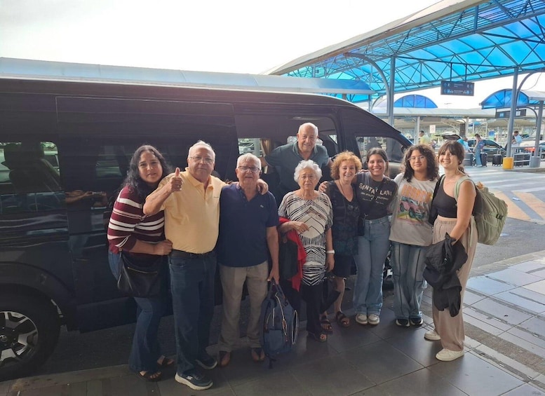 A group of people posing in front of a black van at a bus station with a blue roof structure. 1