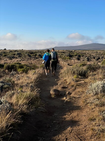 Two people walking on a dirt path surrounded by dry grass and rocks. 2