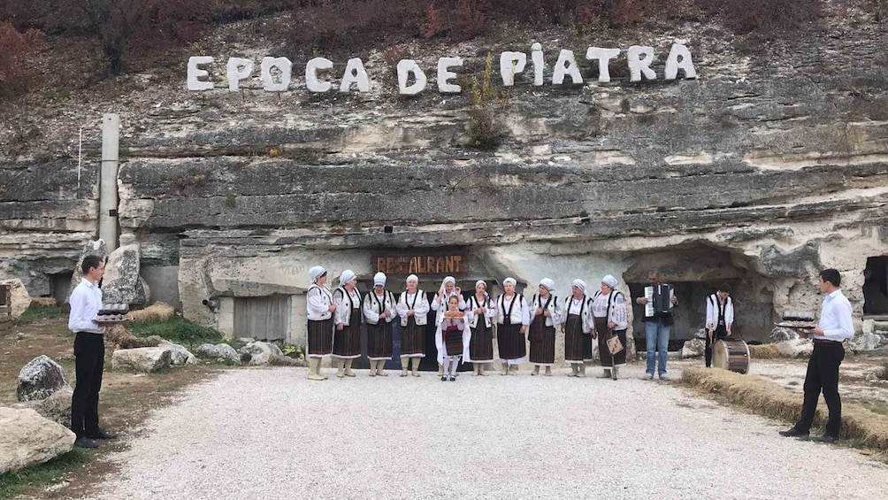 A group of people in traditional attire standing in front of a rock face with the inscription 'EPOCA DE PIATRA'. 2