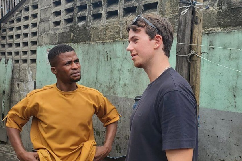 Two individuals standing in an urban setting with a concrete wall and a wooden pole in the background. 3