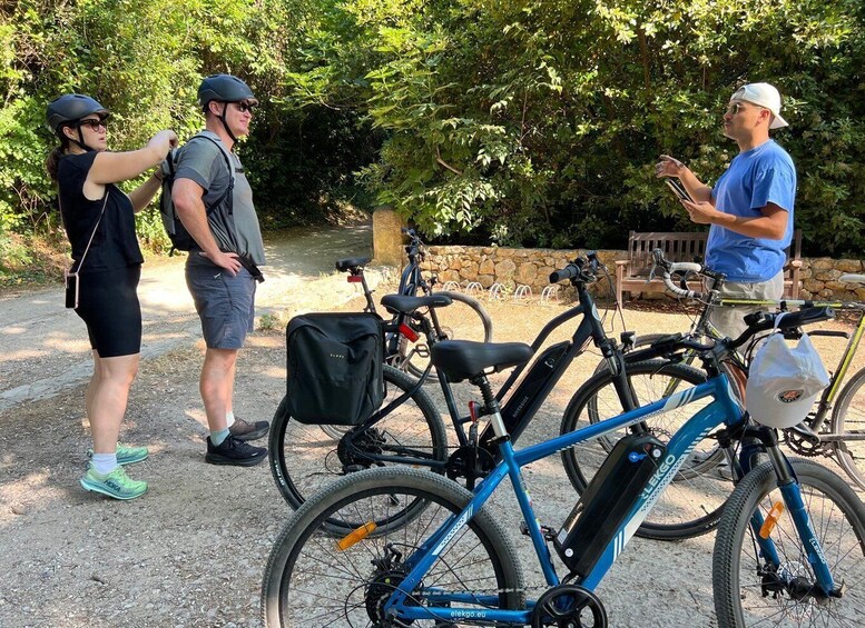 Three people standing near bicycles, with a stone wall and greenery in the background. 2