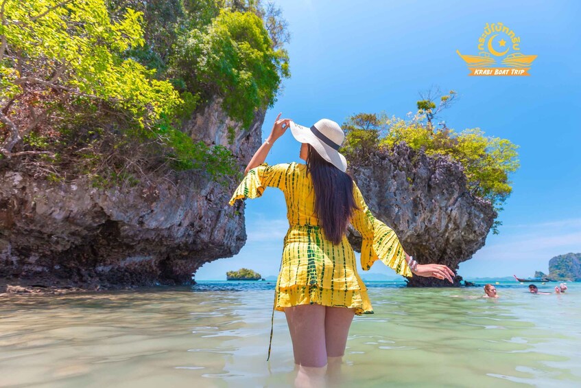 A person in a yellow dress and white hat standing in shallow water near a rocky cliff with greenery, with a clear sky above. 1