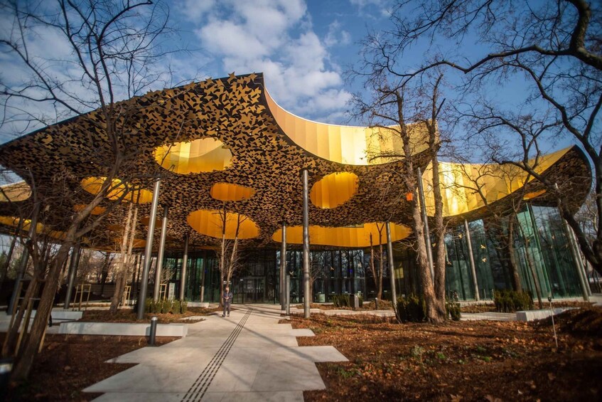 Une structure architecturale moderne, dotée d’un auvent composé de panneaux dorés et de motifs évoquant des feuilles, le tout entouré d’arbres et surplombé par un ciel dégagé. 5
