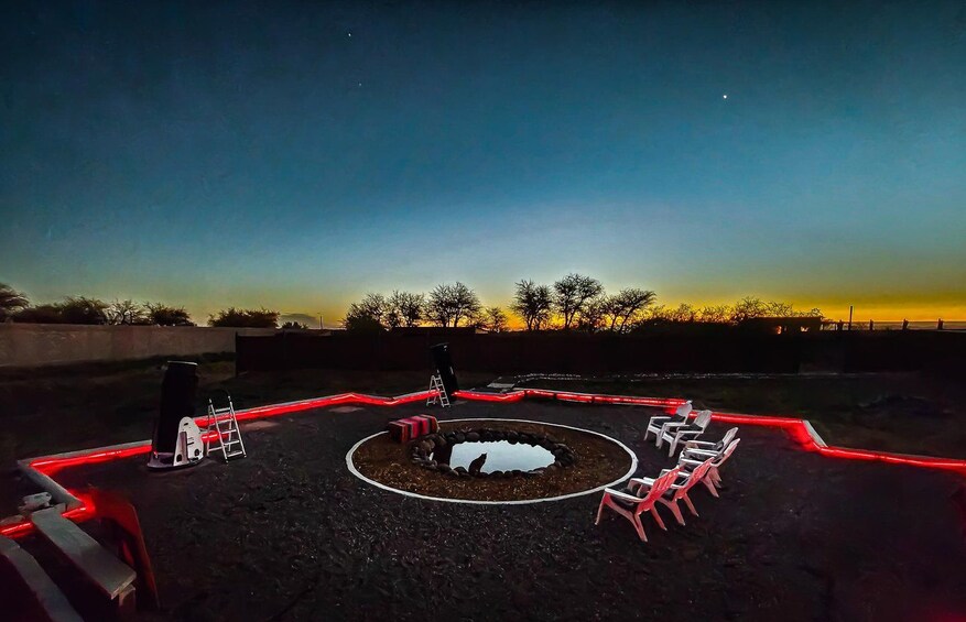 Un área circular con asientos, iluminación roja, una fogata en el centro y árboles silueteados contra un cielo al atardecer. 1