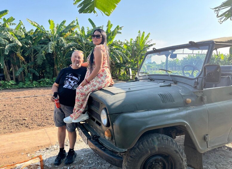 Two people sitting on a green jeep parked in a rural area with banana trees in the background. 5