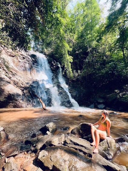 A waterfall in a forested area with a person sitting on rocks by the water. 2