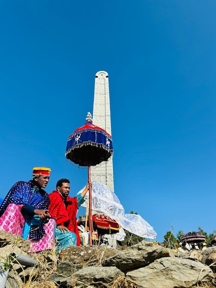 A group of people in colorful traditional attire sitting on rocks with a tall white obelisk in the background. 3