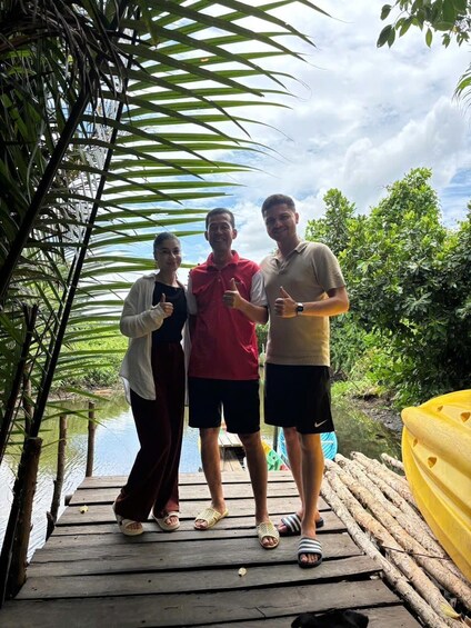 Three people on a wooden dock with a yellow kayak, surrounded by greenery and a body of water. 2