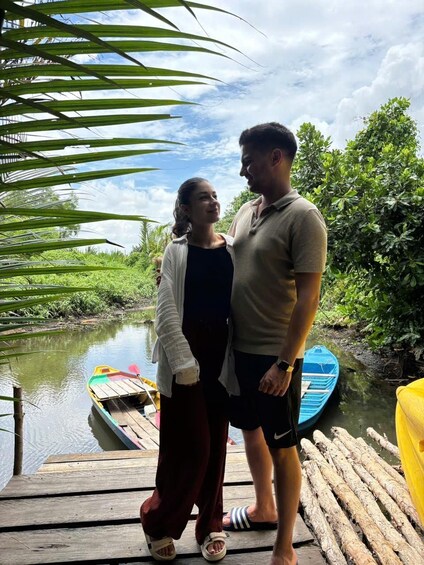 Two people standing on a wooden dock by a calm river, with boats and greenery in the background. 4