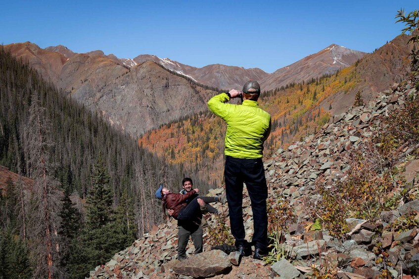 A mountainous landscape with a person taking a photo, another person being carried, and autumn-colored trees. 1