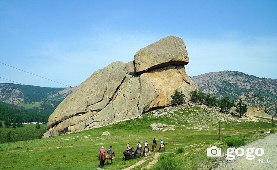A group of people riding horses on a trail with a large rock formation in the background. 5