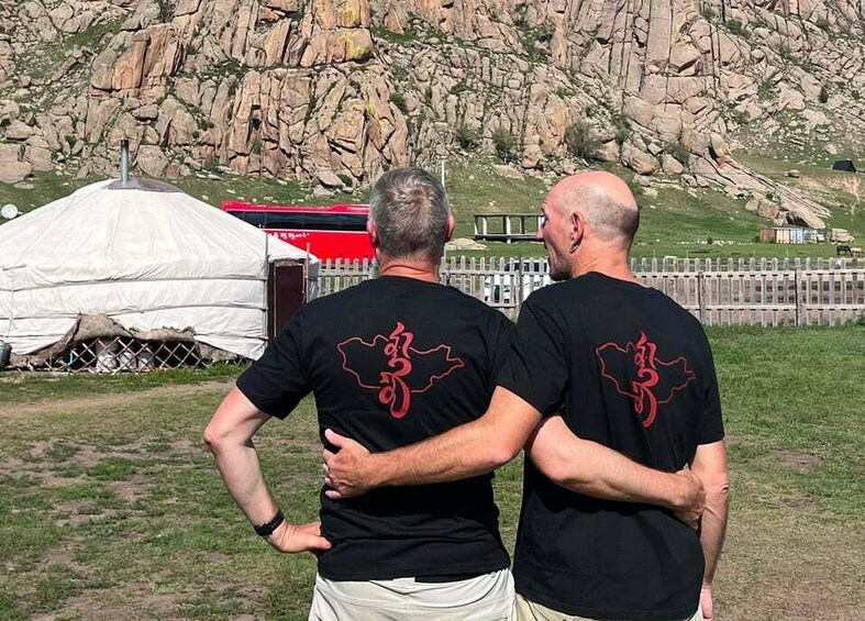 Two people standing with their backs to the camera, wearing black t-shirts with a red design, in front of a rocky landscape with a white tent and a red vehicle in the background. 2