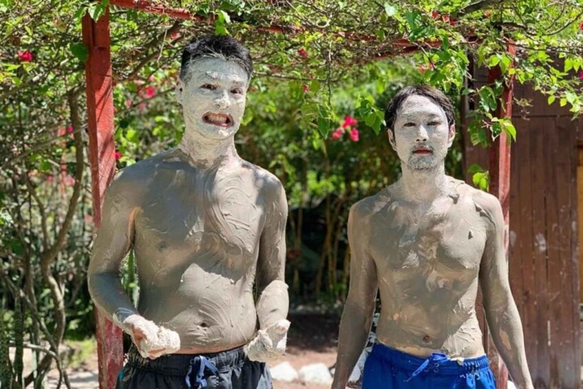 Two shirtless men covered in mud standing under a tree. 1
