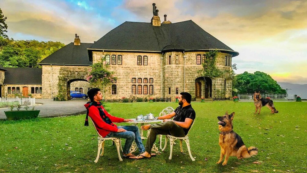 Two people sitting at a table outdoors with a dog in the background. 1