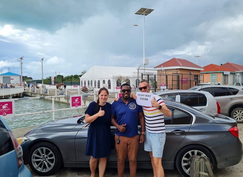Three people posing in front of a car near a waterfront with a white building and a sign that reads "ANTIGUA CRUISE PORT". 2