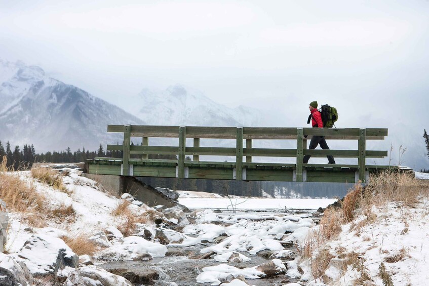 A hiker crossing a wooden bridge over a snow-covered stream with mountains in the background. 1