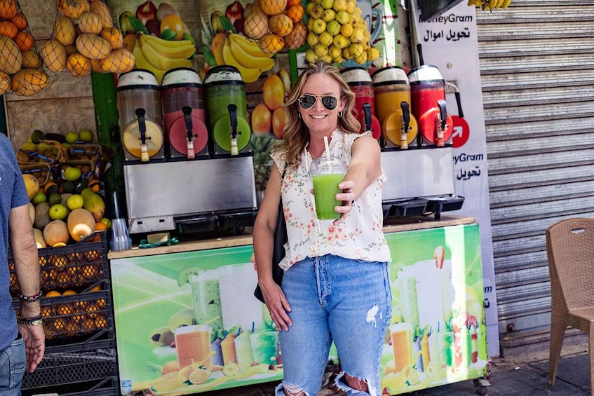 A person holding a green smoothie at a juice stand with various fruits and juicers in the background. 2